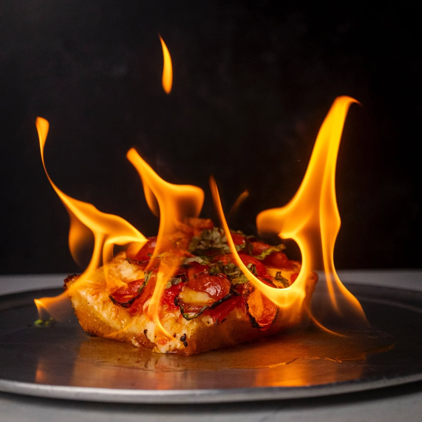 Square pepperoni slice with basil on a pan, surrounded by open flames, shot for Pizza One