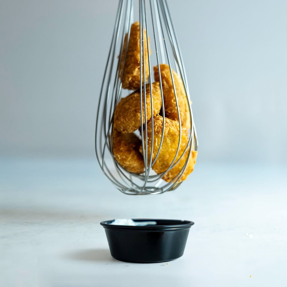 Fried chicken pieces held in a wire whisk above a black cup of dipping sauce